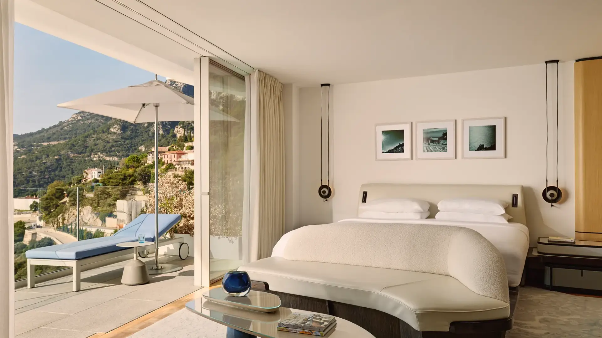 Chambre d’hôtel élégante avec grand lit, banquette incurvée et portes vitrées s’ouvrant sur une terrasse avec transat, parasol et vue sur la montagne.