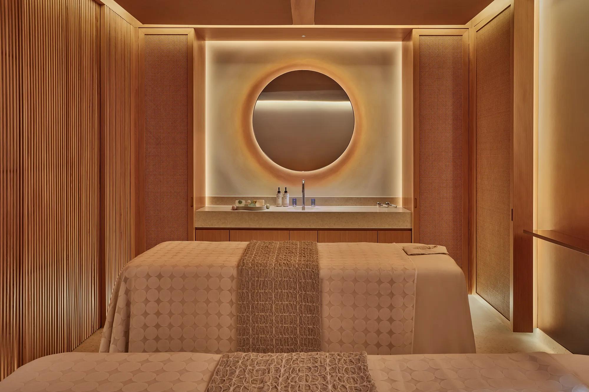 Warm-toned spa treatment room with twin beds, circular backlit mirror, and minimalist wood-paneled walls
