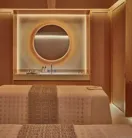 Warm-toned spa treatment room with twin beds, circular backlit mirror, and minimalist wood-paneled walls