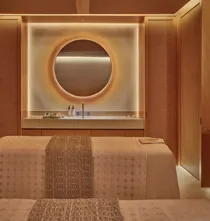 Warm-toned spa treatment room with twin beds, circular backlit mirror, and minimalist wood-paneled walls