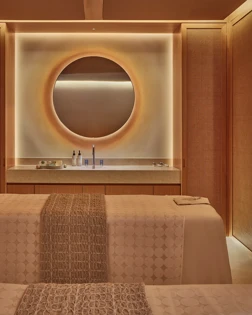 Warm-toned spa treatment room with twin beds, circular backlit mirror, and minimalist wood-paneled walls