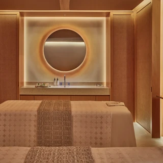 Warm-toned spa treatment room with twin beds, circular backlit mirror, and minimalist wood-paneled walls