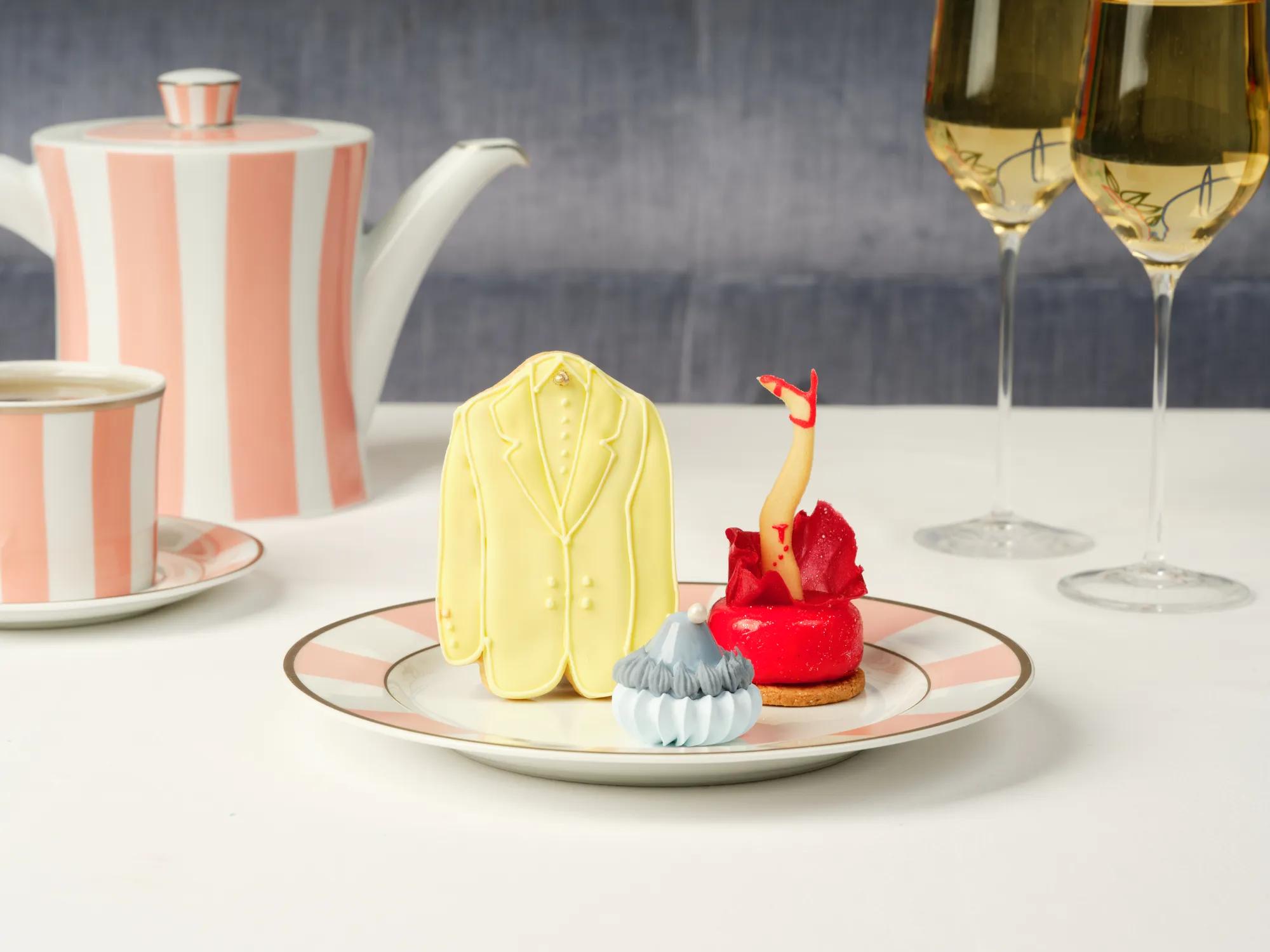 A yellow suit cookie, red leg pastry, and blue meringue treat on a plate with striped tea china behind.