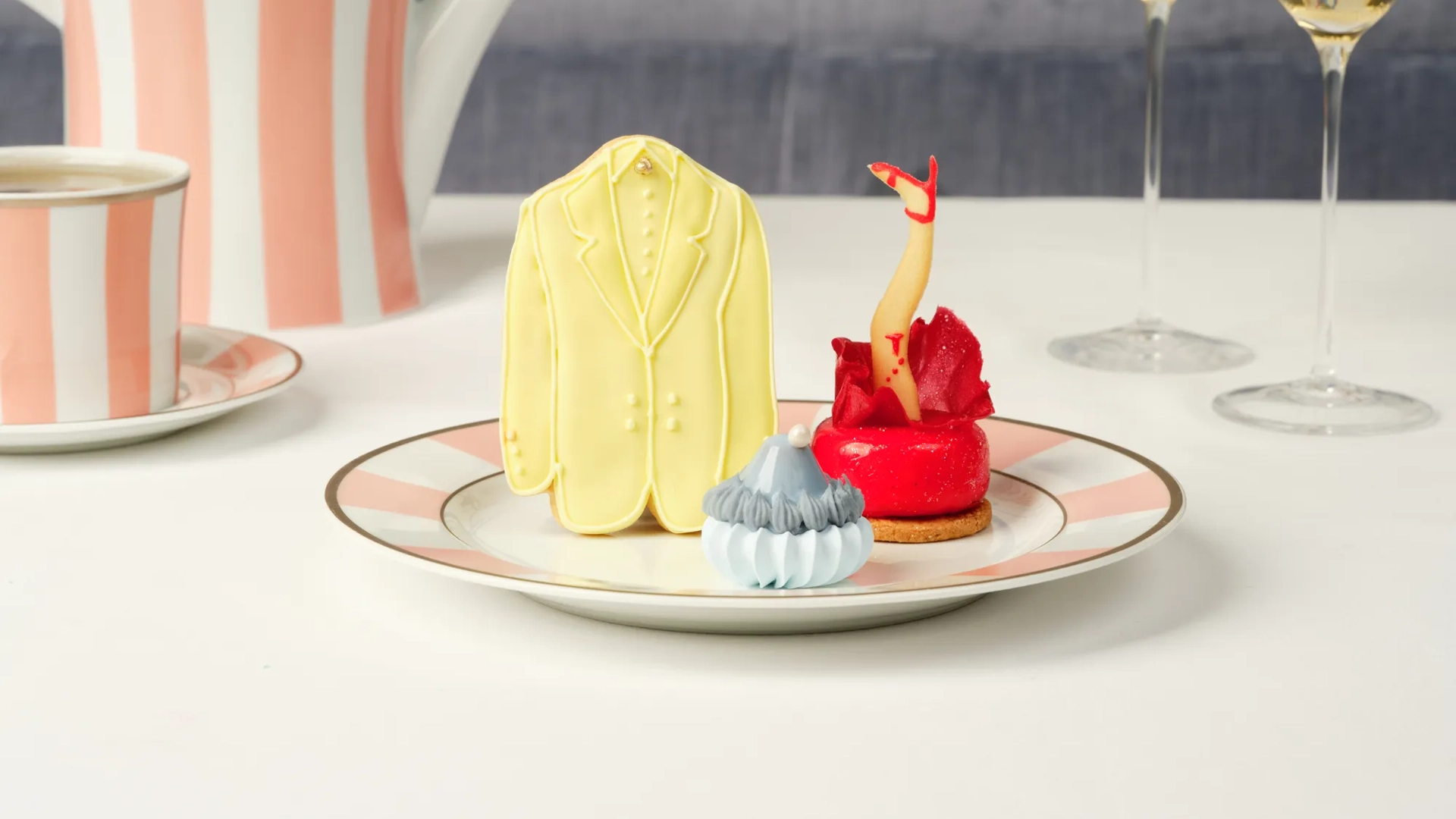 A yellow suit cookie, red leg pastry, and blue meringue treat on a plate with striped tea china behind.