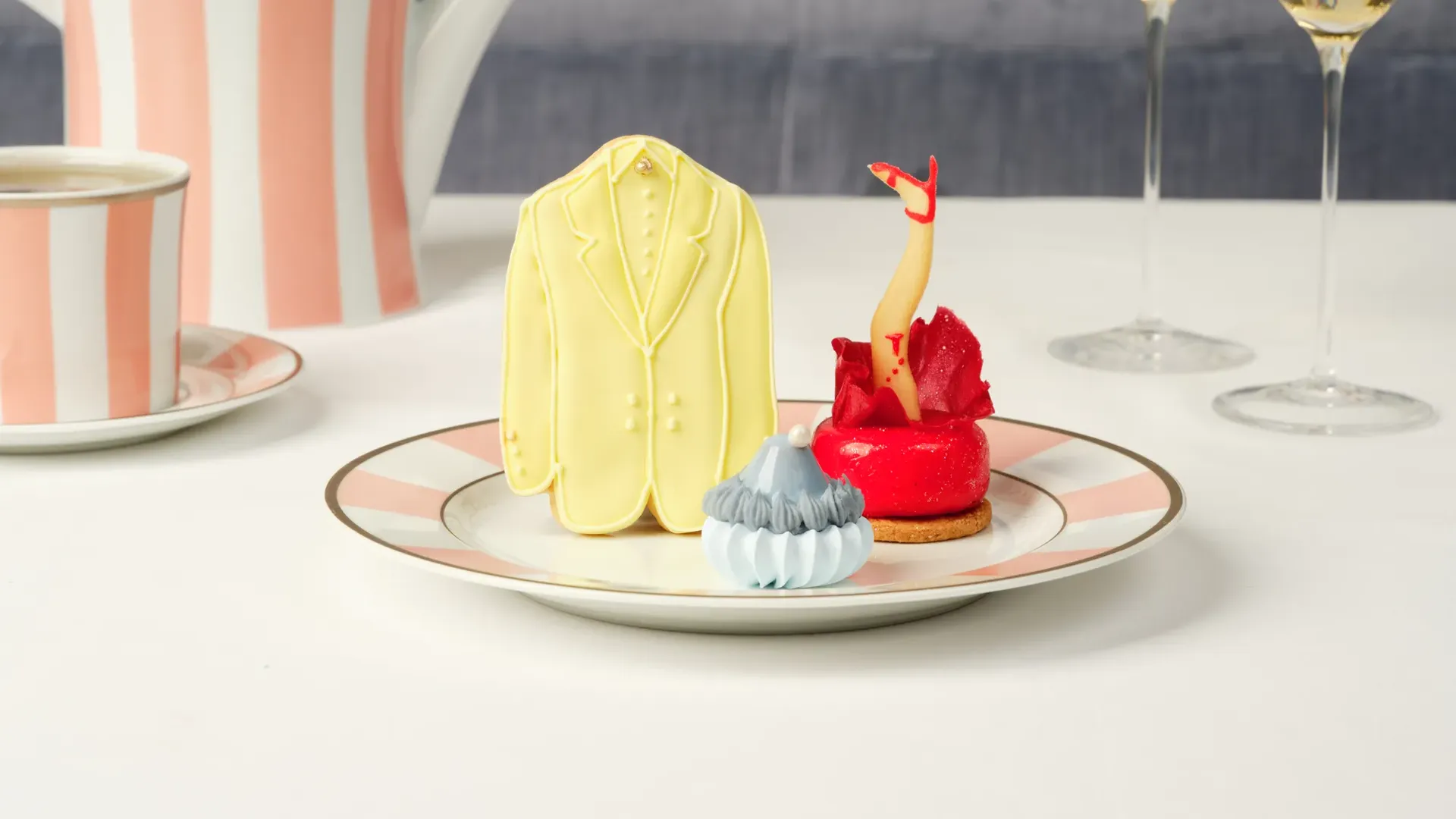 A yellow suit cookie, red leg pastry, and blue meringue treat on a plate with striped tea china behind.