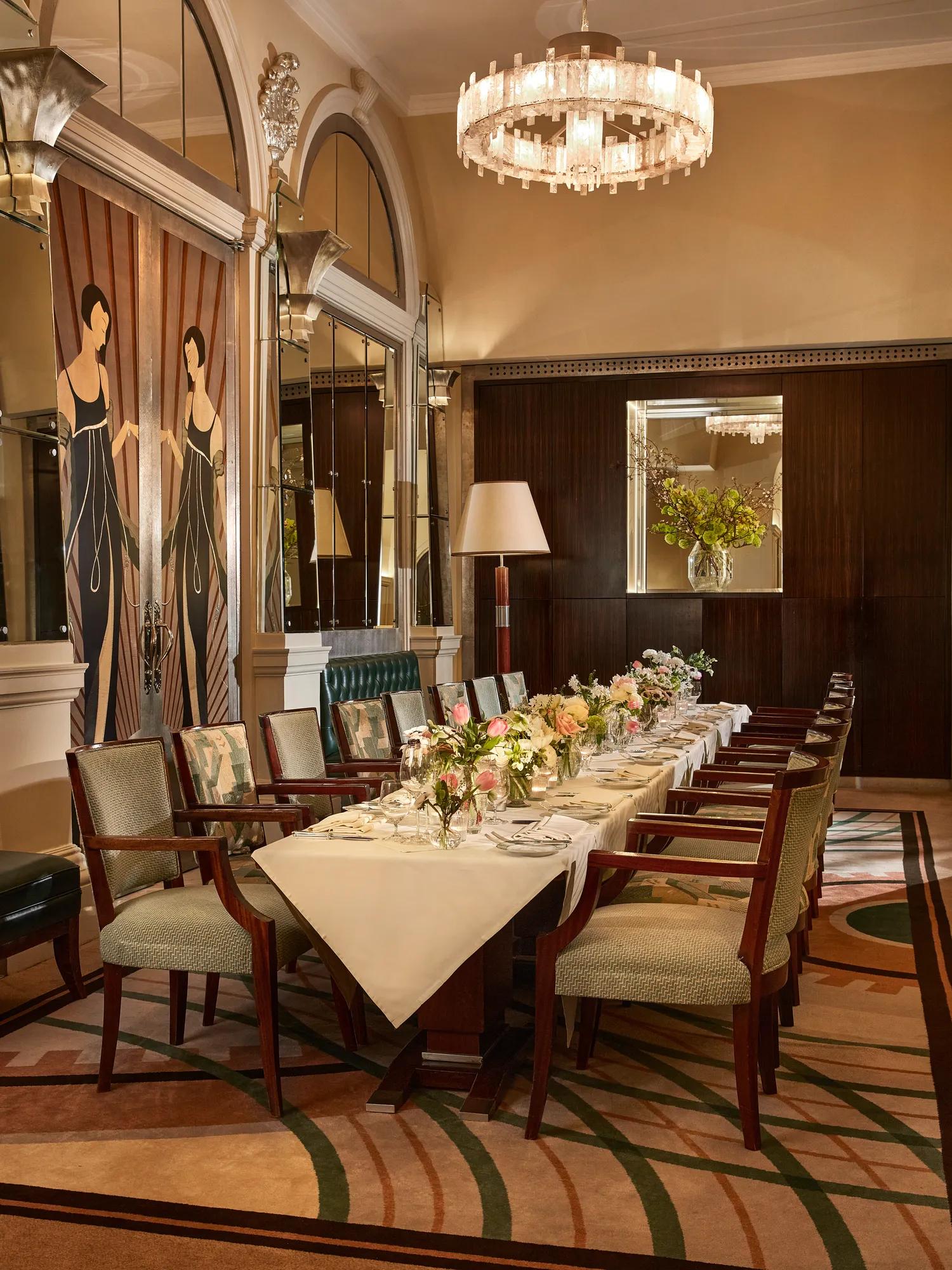 Elegant private dining room with an Art Deco design, featuring a long table set with white linens, floral arrangements, and upholstered chairs beneath a crystal chandelier.