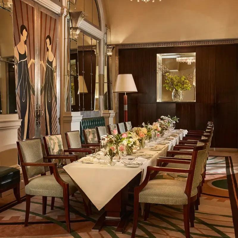Elegant private dining room with an Art Deco design, featuring a long table set with white linens, floral arrangements, and upholstered chairs beneath a crystal chandelier.