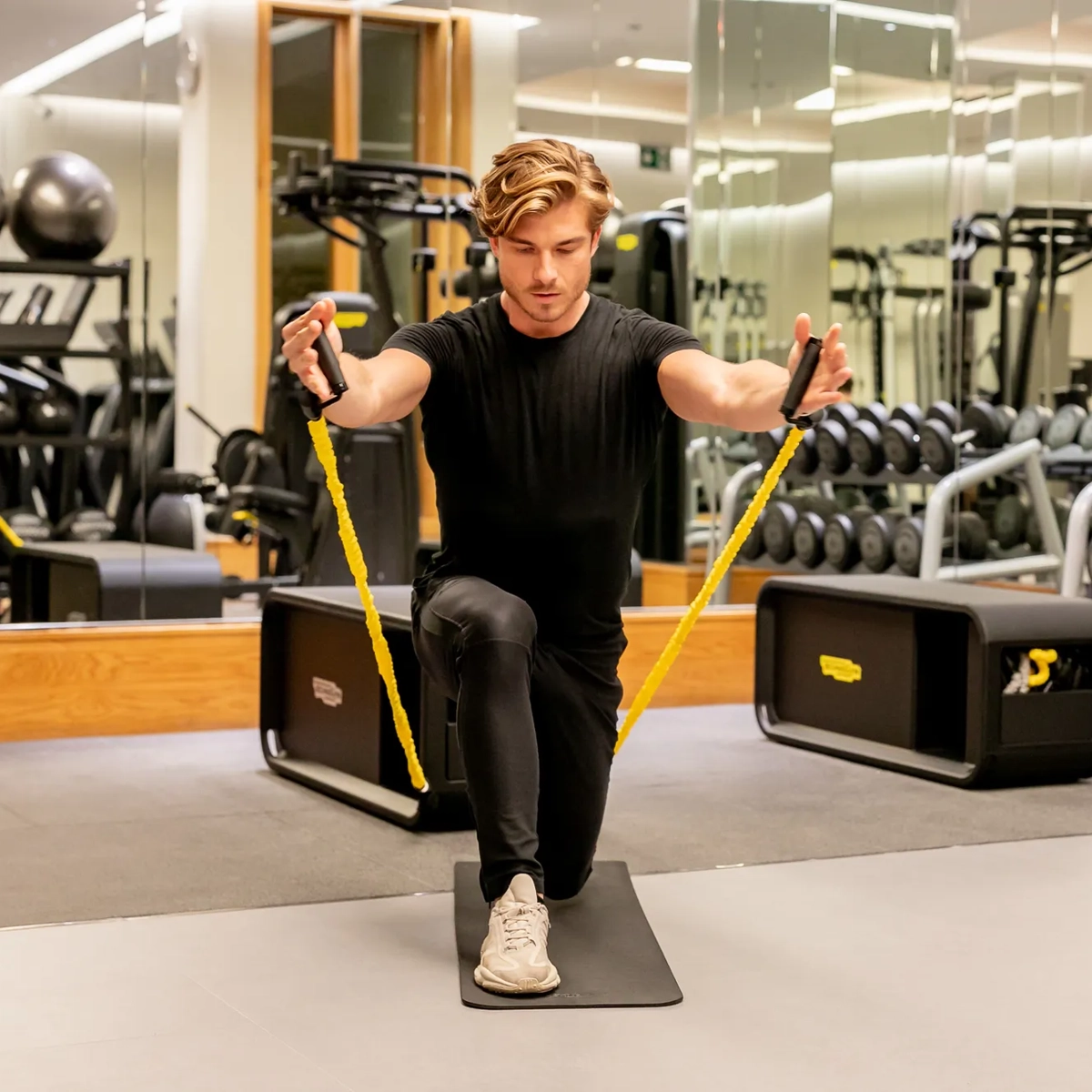 Man kneeling on a gym mat using yellow resistance bands for an upper-body exercise.