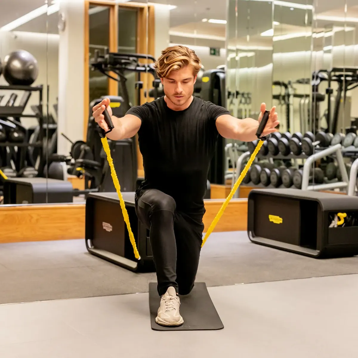 Man kneeling on a gym mat using yellow resistance bands for an upper-body exercise.