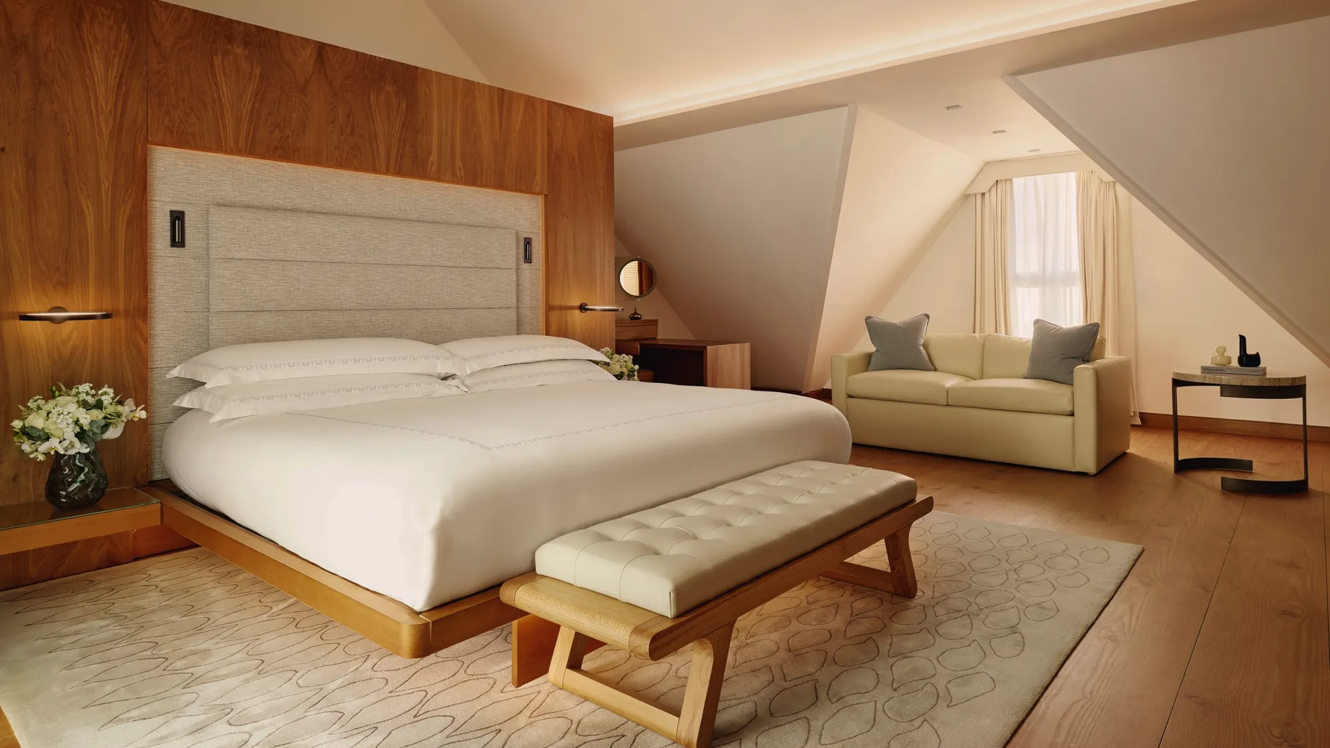 Serene attic bedroom with wooden feature wall, upholstered bed, cream sofa, and bench on a soft rug.