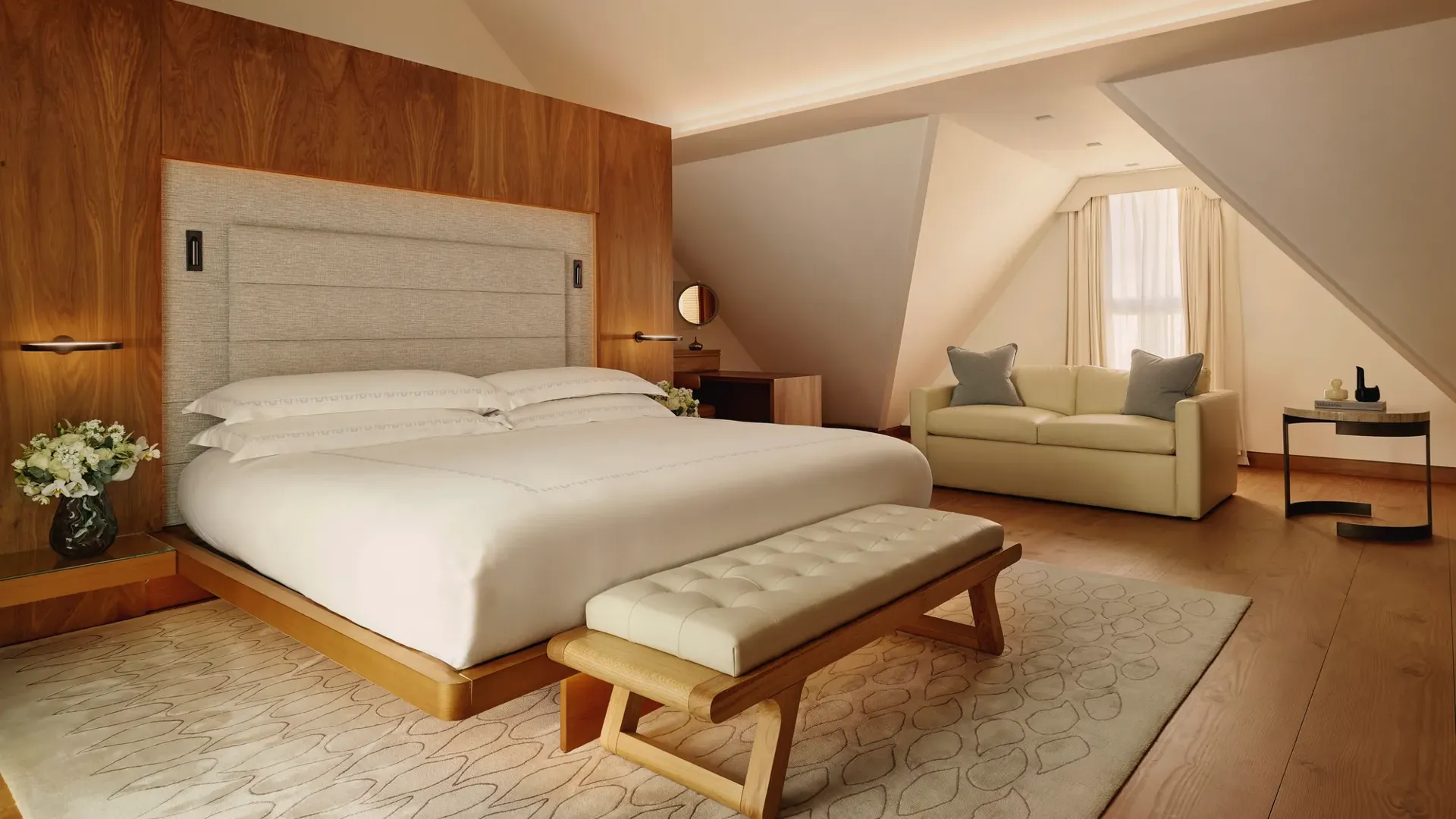 Serene attic bedroom with wooden feature wall, upholstered bed, cream sofa, and bench on a soft rug.