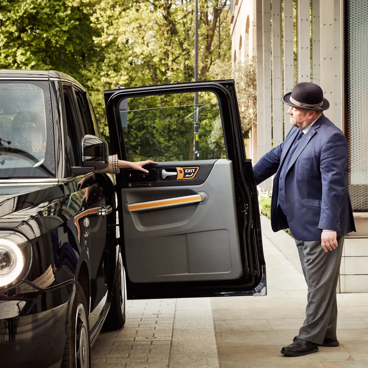 A doorman in a navy suit holds open the door of a black taxi as a guest steps out at the hotel entrance.
