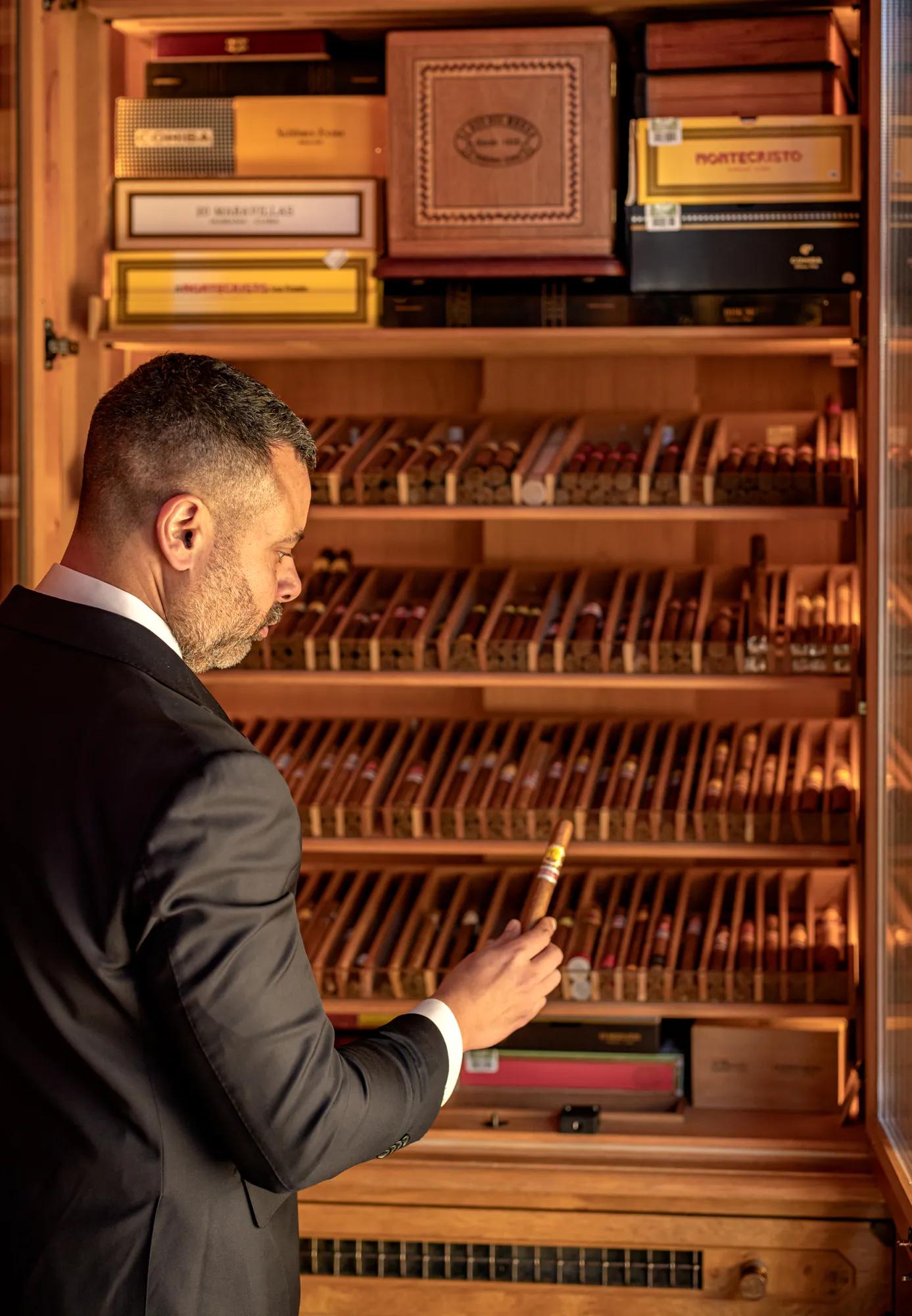 Man in suit selecting a cigar from a well-stocked wooden humidor cabinet