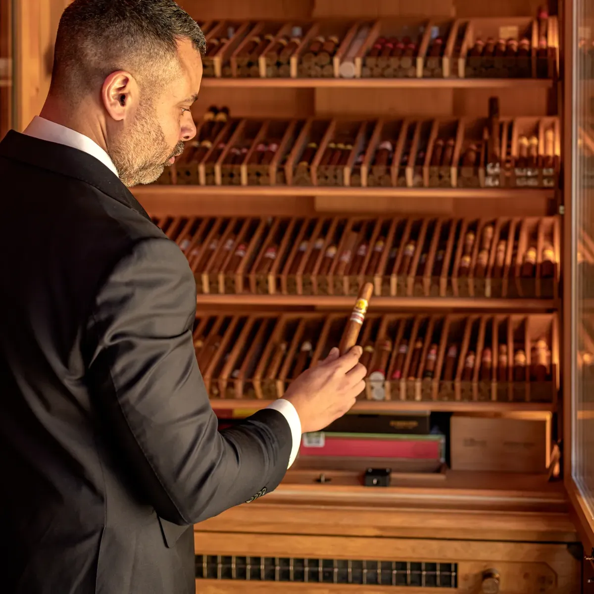 Man in suit selecting a cigar from a well-stocked wooden humidor cabinet