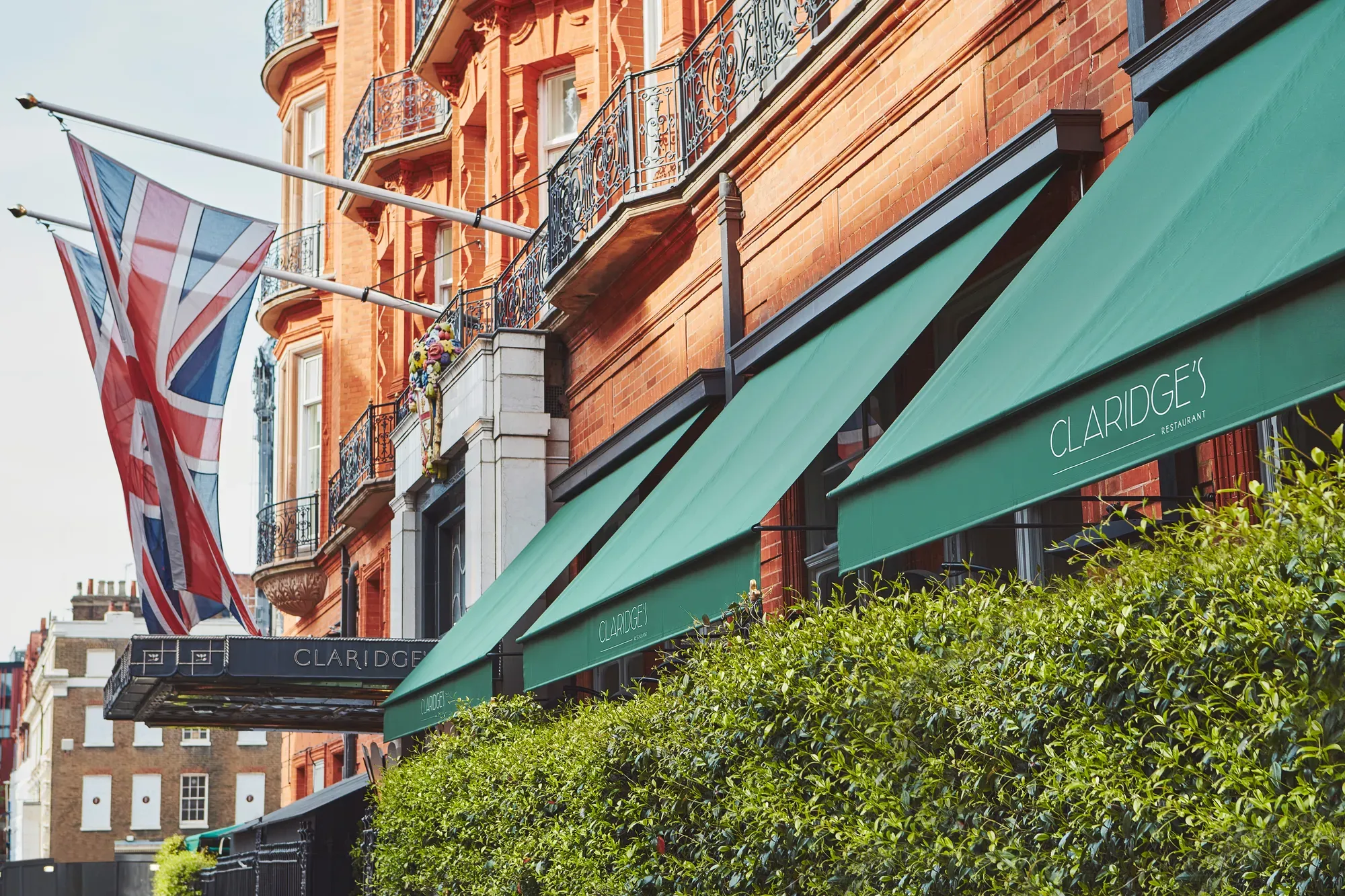 Red-brick hotel façade with green Claridge’s awnings, Union Jack flags and wrought-iron balconies above leafy hedge