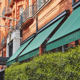 Red-brick hotel façade with green Claridge’s awnings, Union Jack flags and wrought-iron balconies above leafy hedge
