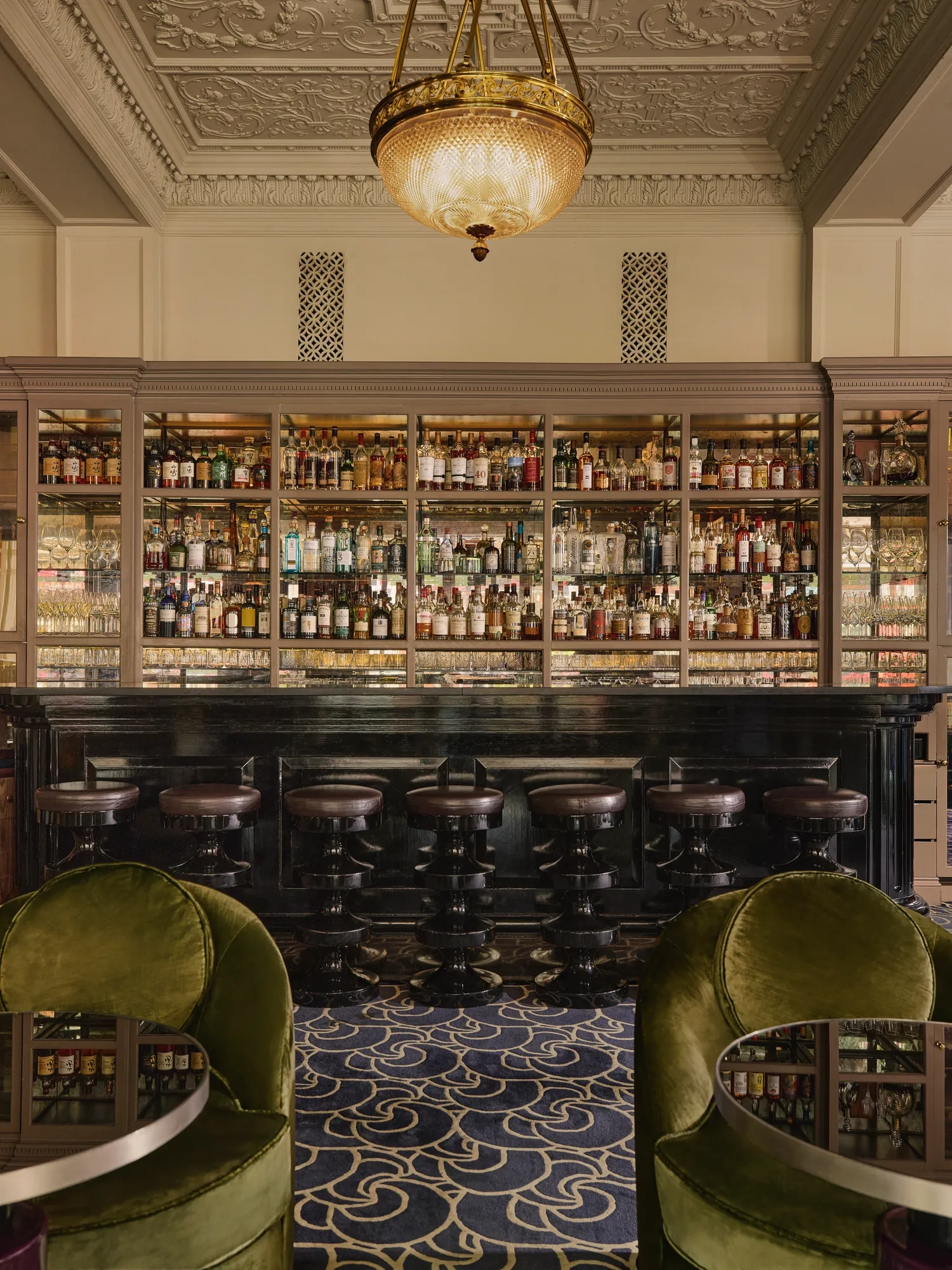 Elegant bar with a dark counter, high stools, mirrored shelves of spirits, and green velvet lounge chairs beneath a chandelier.