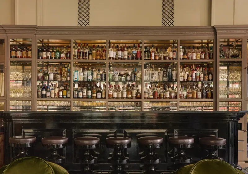 Elegant bar with a dark counter, high stools, mirrored shelves of spirits, and green velvet lounge chairs beneath a chandelier.