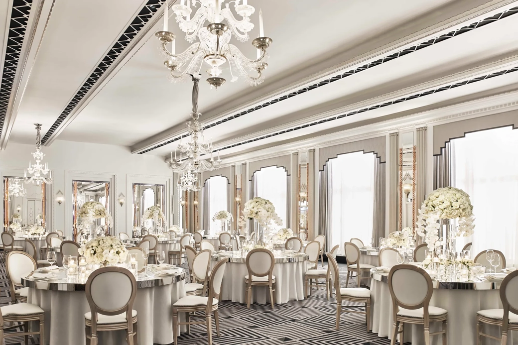 The Ballroom at Claridge’s set for an elegant event with round tables draped in white linens, floral centrepieces of white blooms, crystal chandeliers, and mirrored Art Deco details.
