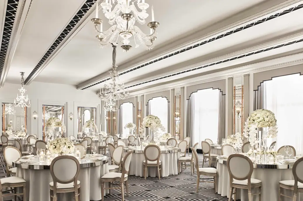 The Ballroom at Claridge’s set for an elegant event with round tables draped in white linens, floral centrepieces of white blooms, crystal chandeliers, and mirrored Art Deco details.