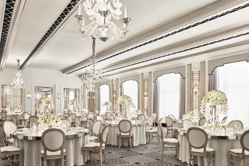 The Ballroom at Claridge’s set for an elegant event with round tables draped in white linens, floral centrepieces of white blooms, crystal chandeliers, and mirrored Art Deco details.
