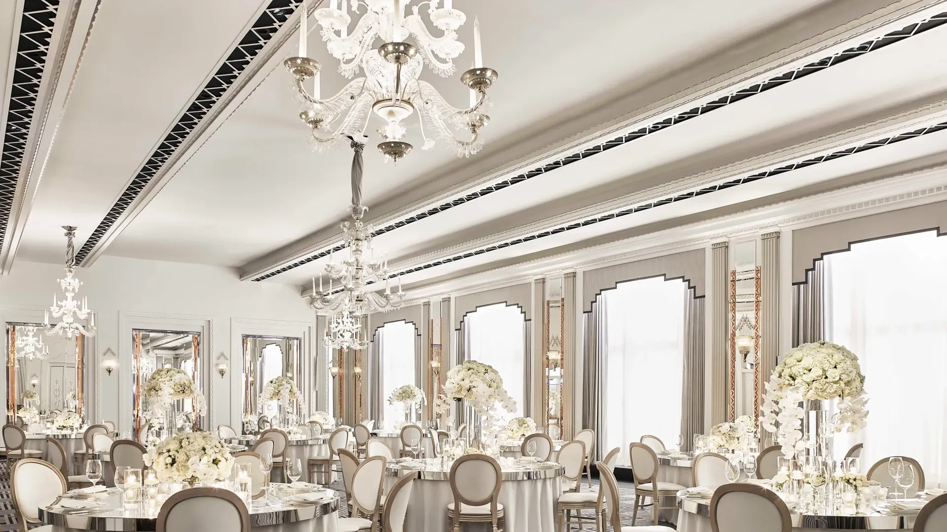 The Ballroom at Claridge’s set for an elegant event with round tables draped in white linens, floral centrepieces of white blooms, crystal chandeliers, and mirrored Art Deco details.