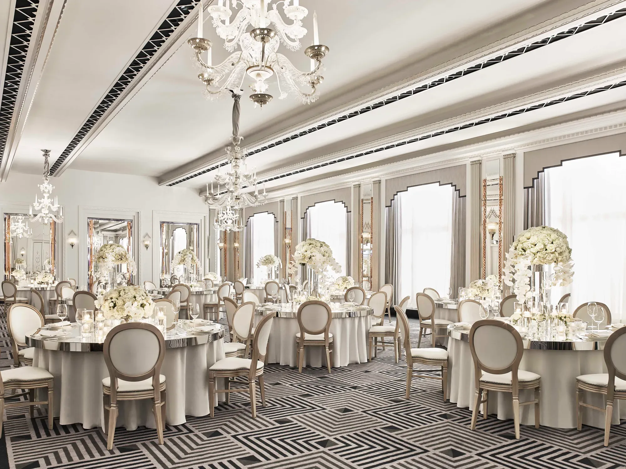 The Ballroom at Claridge’s set for an elegant event with round tables draped in white linens, floral centrepieces of white blooms, crystal chandeliers, and mirrored Art Deco details.