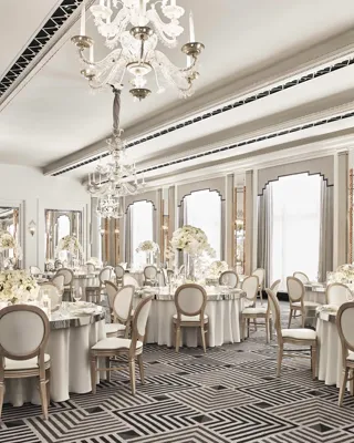 The Ballroom at Claridge’s set for an elegant event with round tables draped in white linens, floral centrepieces of white blooms, crystal chandeliers, and mirrored Art Deco details.