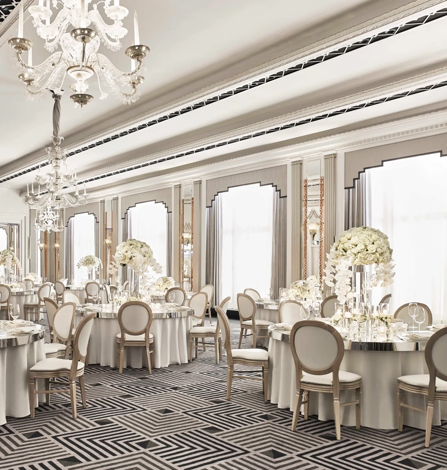 The Ballroom at Claridge’s set for an elegant event with round tables draped in white linens, floral centrepieces of white blooms, crystal chandeliers, and mirrored Art Deco details.