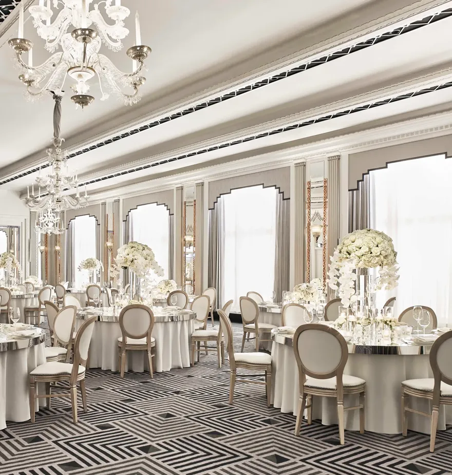 The Ballroom at Claridge’s set for an elegant event with round tables draped in white linens, floral centrepieces of white blooms, crystal chandeliers, and mirrored Art Deco details.