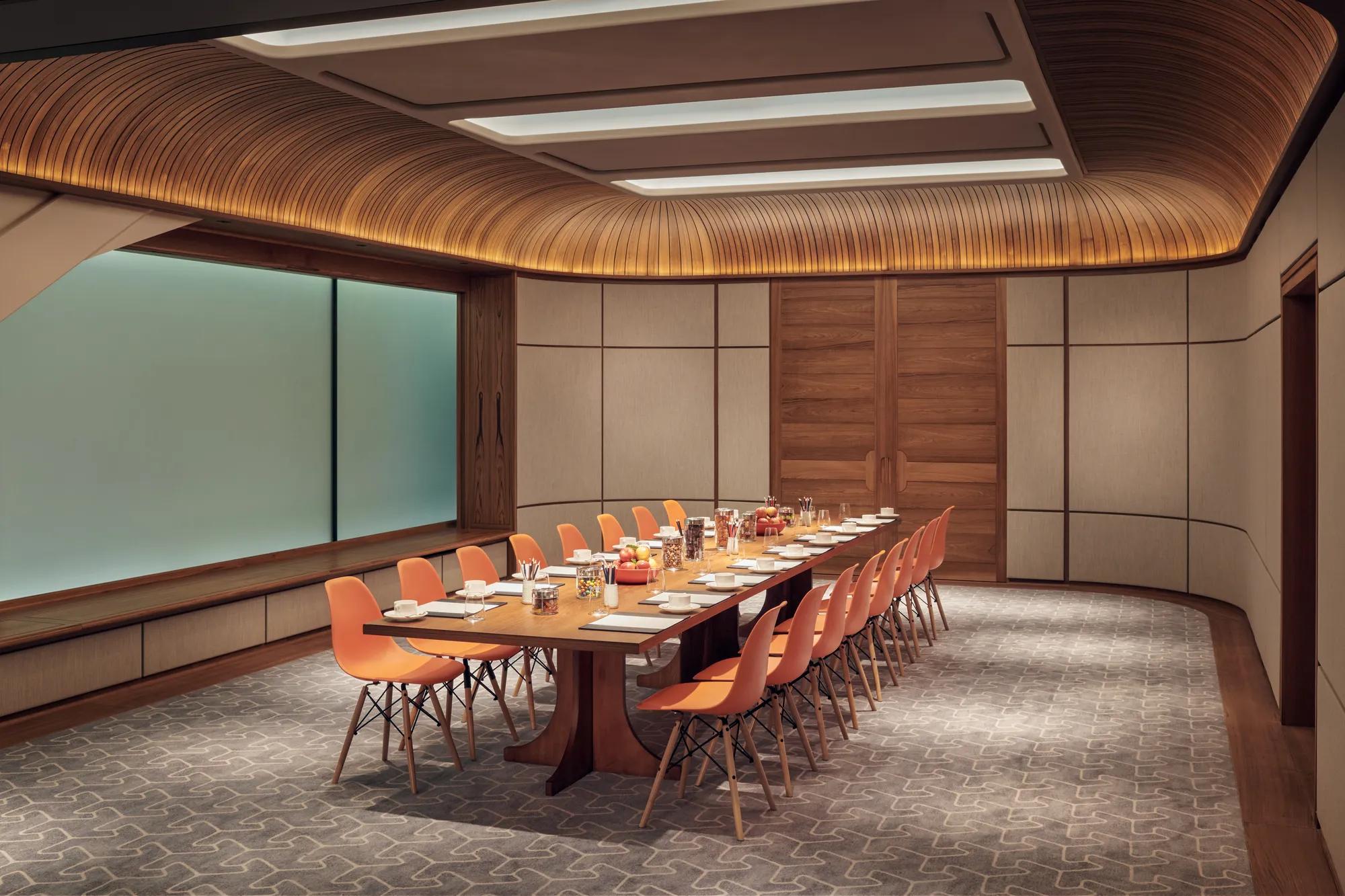 Modern meeting room with long wooden table, orange chairs, and wood-panelled ceiling set for a business event.