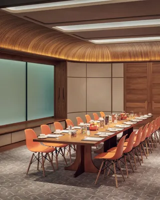Modern meeting room with long wooden table, orange chairs, and wood-panelled ceiling set for a business event.