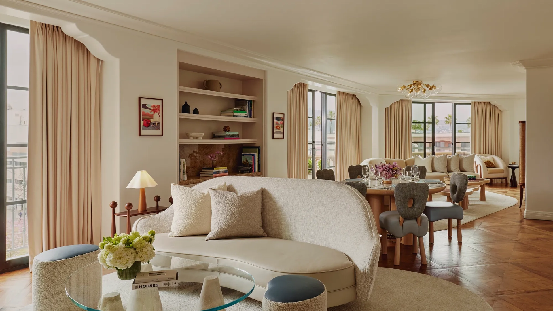 Expansive suite interior: sculptural glass coffee table and curved sofa in foreground; round dining table set with florals; built‑in shelves; large draped windows.