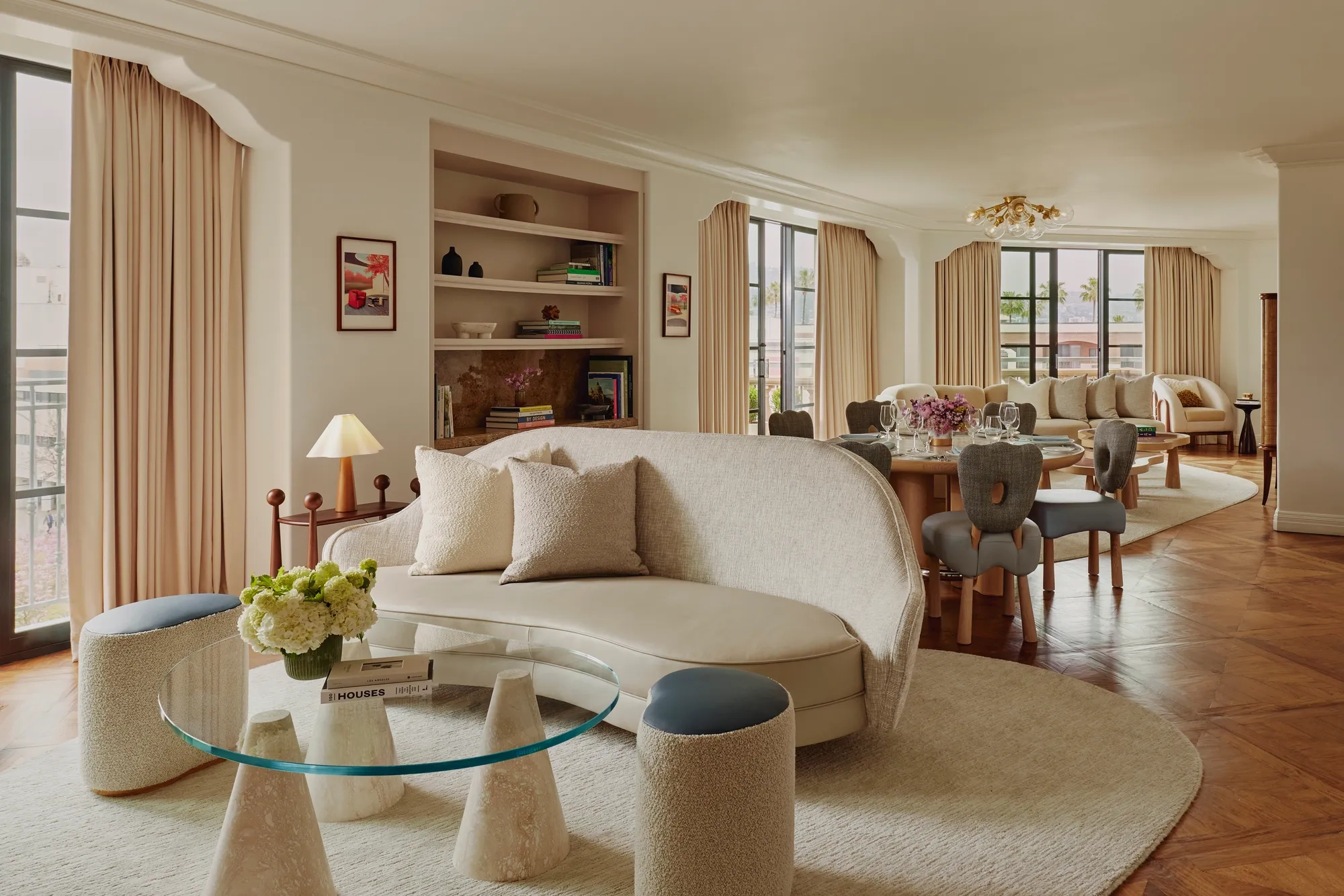 Expansive suite interior: sculptural glass coffee table and curved sofa in foreground; round dining table set with florals; built‑in shelves; large draped windows.