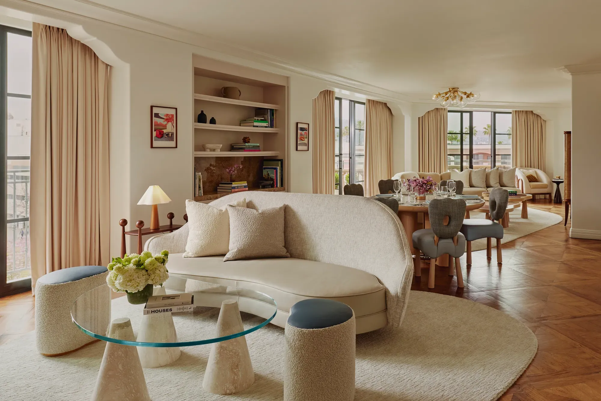 Expansive suite interior: sculptural glass coffee table and curved sofa in foreground; round dining table set with florals; built‑in shelves; large draped windows.