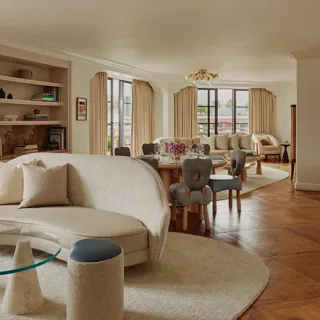 Expansive suite interior: sculptural glass coffee table and curved sofa in foreground; round dining table set with florals; built‑in shelves; large draped windows.