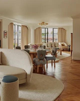 Expansive suite interior: sculptural glass coffee table and curved sofa in foreground; round dining table set with florals; built‑in shelves; large draped windows.