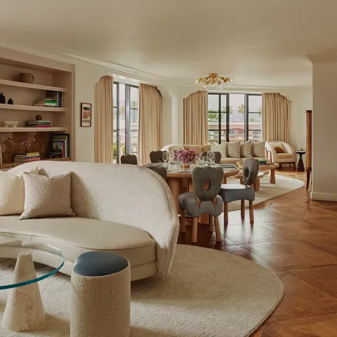 Expansive suite interior: sculptural glass coffee table and curved sofa in foreground; round dining table set with florals; built‑in shelves; large draped windows.