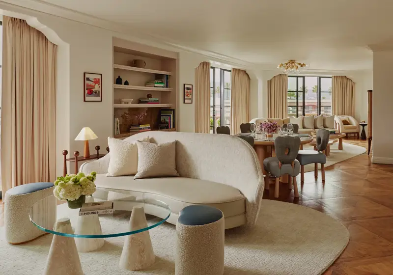 Expansive suite interior: sculptural glass coffee table and curved sofa in foreground; round dining table set with florals; built‑in shelves; large draped windows.