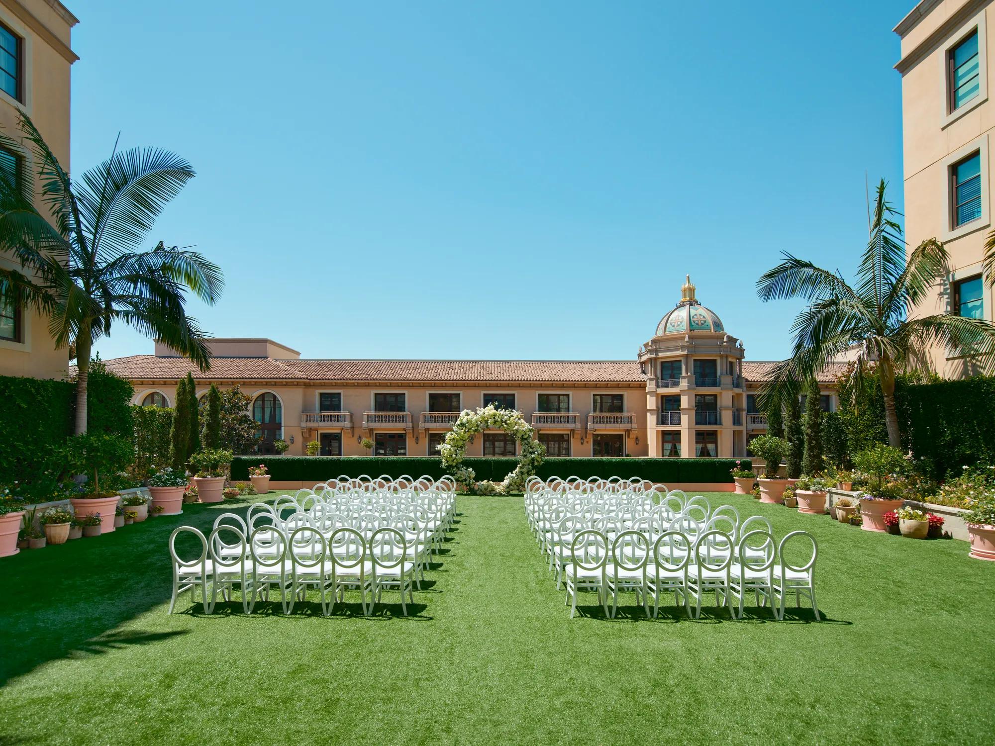 Symmetrical ground-level aisle perspective; petal-lined path between circular-back chairs toward a white floral arch and domed tower.