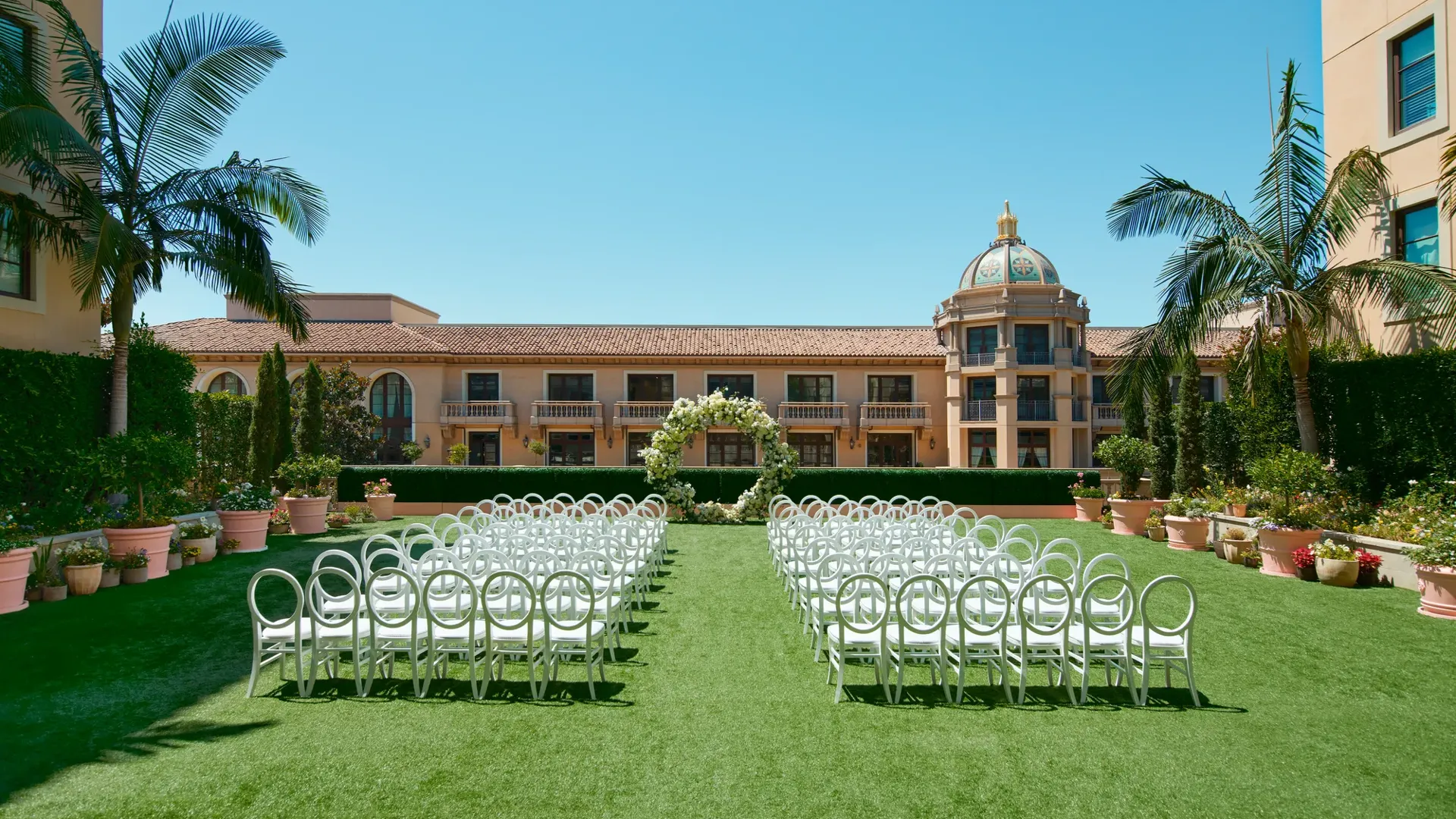 Symmetrical ground-level aisle perspective; petal-lined path between circular-back chairs toward a white floral arch and domed tower.