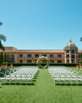 Symmetrical ground-level aisle perspective; petal-lined path between circular-back chairs toward a white floral arch and domed tower.
