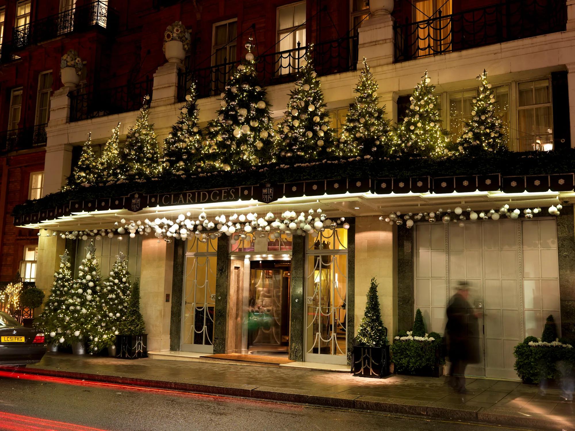 Hotel entrance at night decorated with illuminated Christmas trees and garlands, with canopy lights, revolving door, and blurred pedestrians passing by.