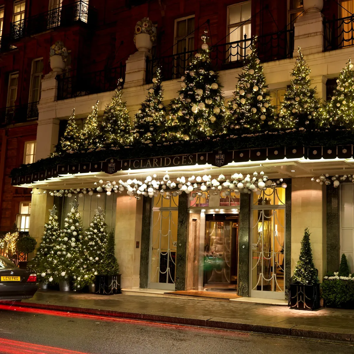 Hotel entrance at night decorated with illuminated Christmas trees and garlands, with canopy lights, revolving door, and blurred pedestrians passing by.