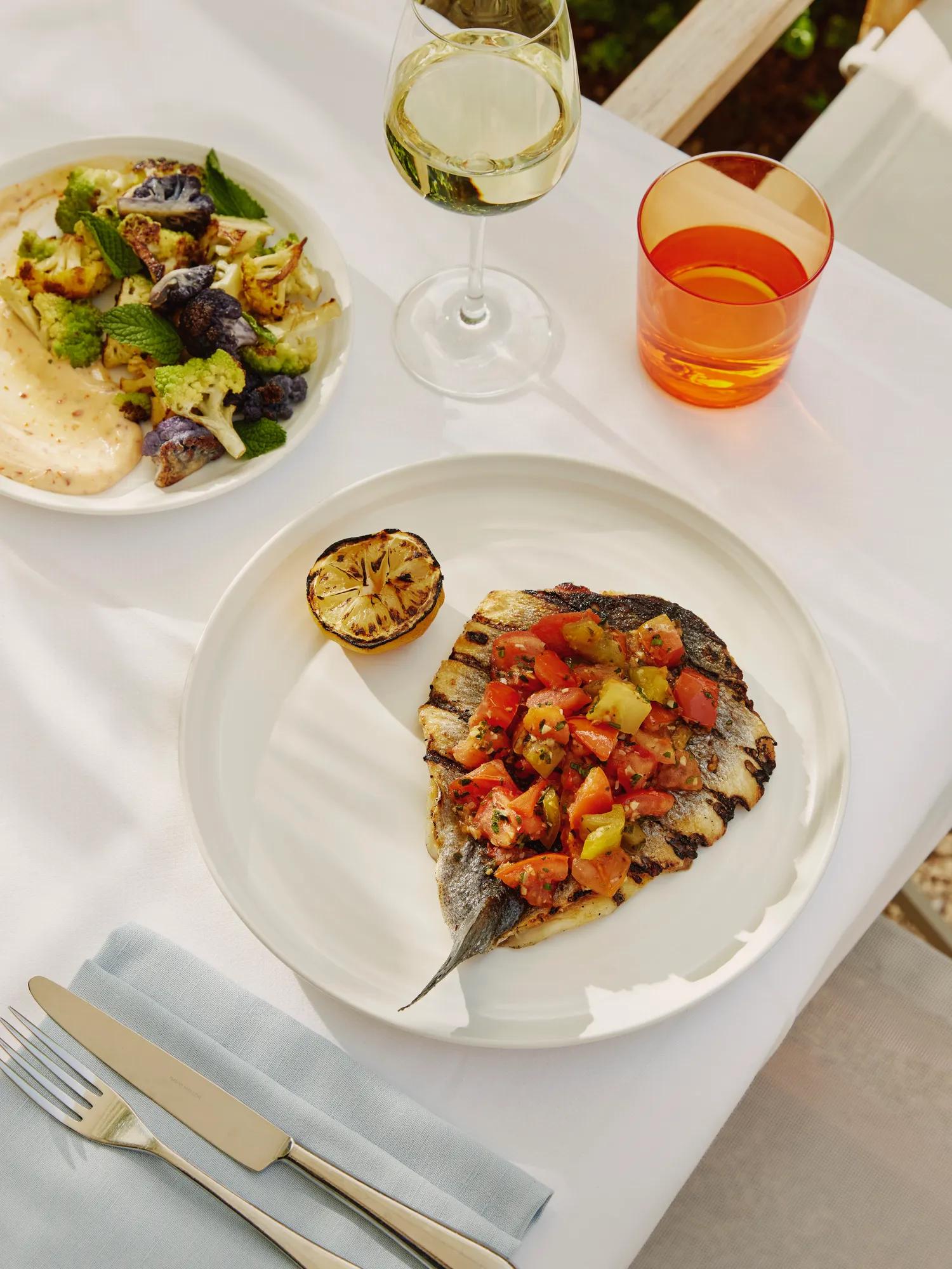 Grilled fish topped with tomato relish and charred lemon; cauliflower side and wine on terrace table.