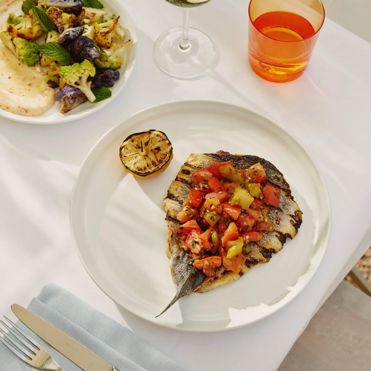 Grilled fish topped with tomato relish and charred lemon; cauliflower side and wine on terrace table.