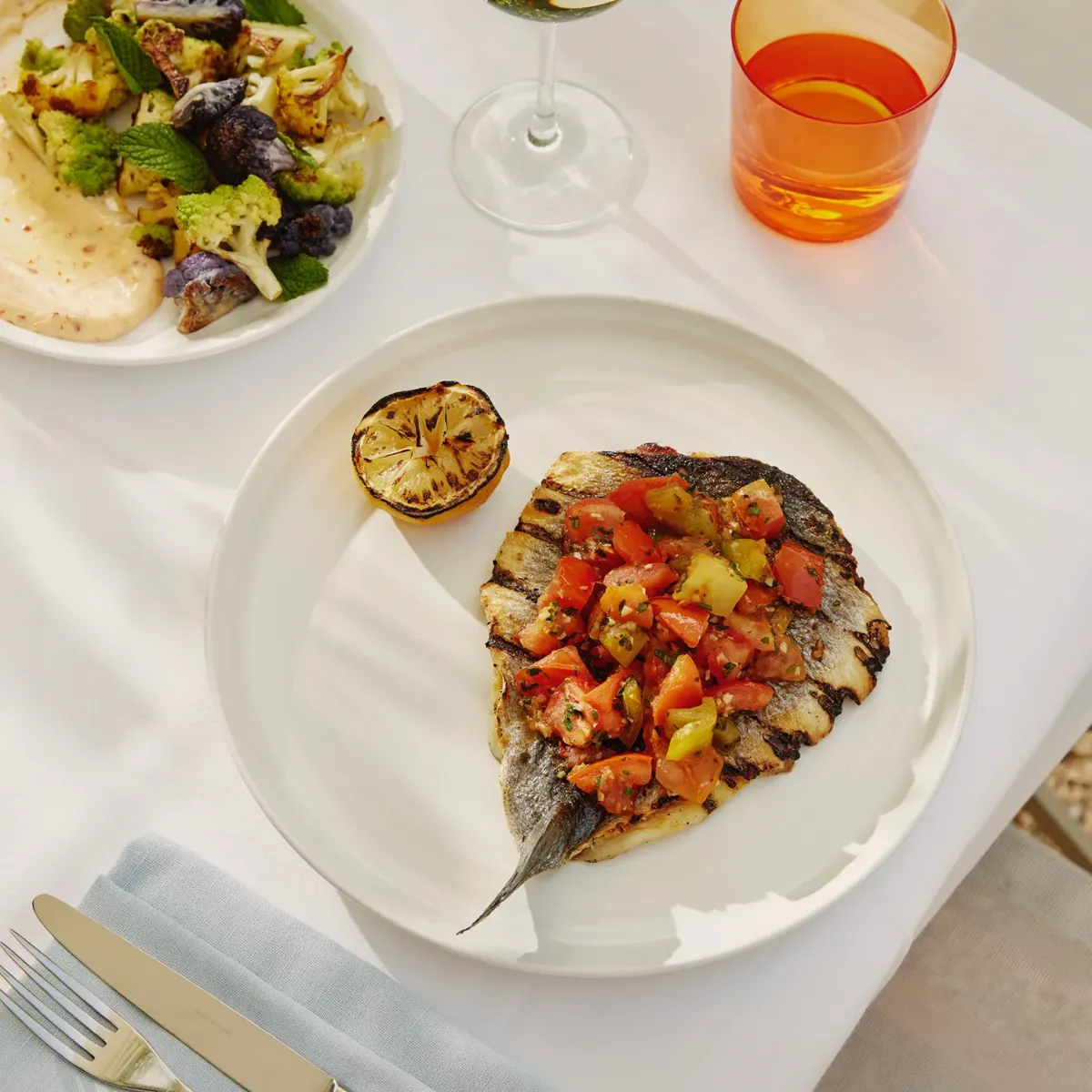 Grilled fish topped with tomato relish and charred lemon; cauliflower side and wine on terrace table.