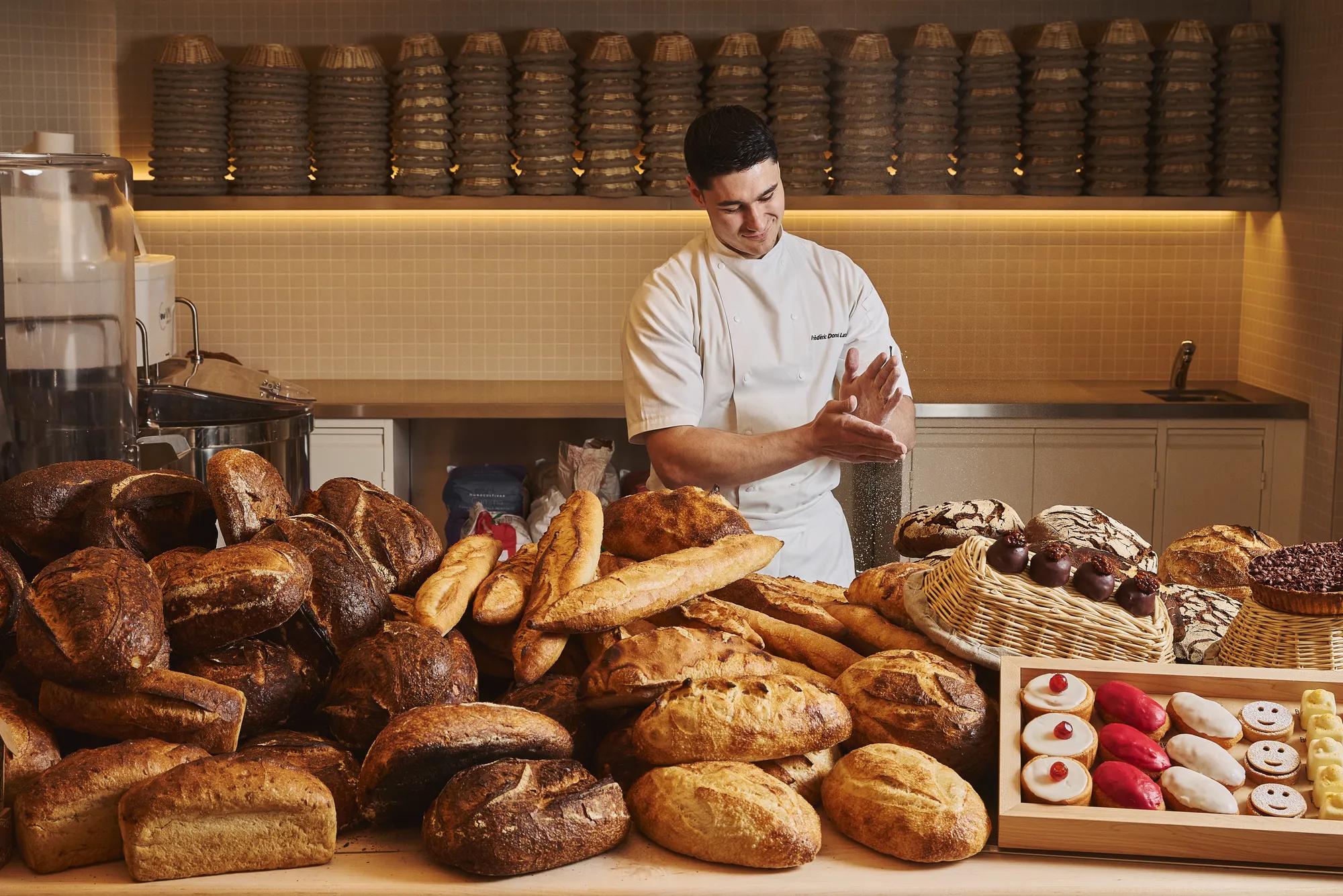 Chef in white uniform arranging loaves and pastries behind large display of assorted breads in bakery kitchen.