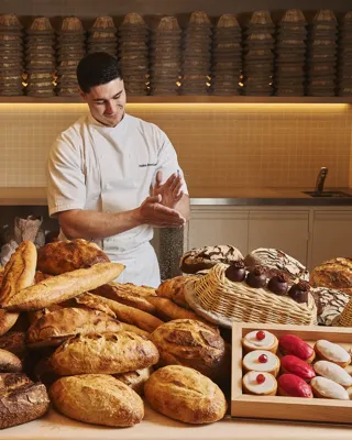 Chef in white uniform arranging loaves and pastries behind large display of assorted breads in bakery kitchen.