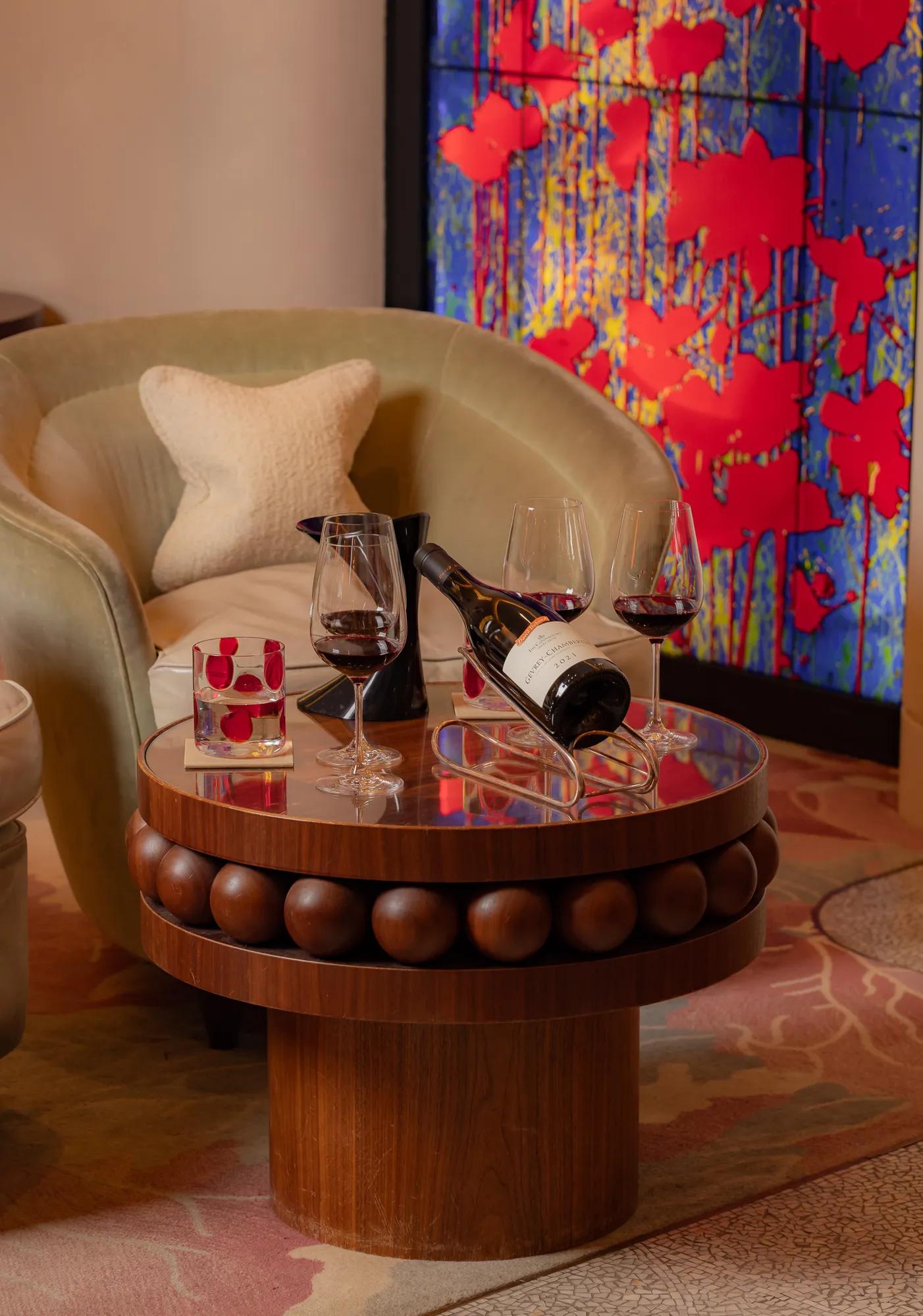 Wine bottle and two glasses on a round table beside an armchair and colourful artwork.